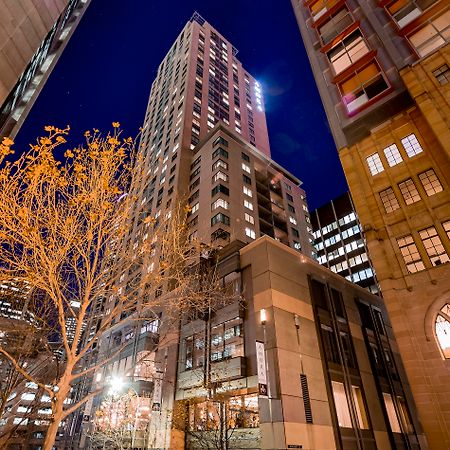 Amora Hotel Jamison Sydney Kültér fotó The photo shows a tall building illuminated at night, surrounded by other structures in an urban environment. The foreground features a tree with bare branches, suggesting it's possibly autumn or winter. The building itself has a modern design, with 