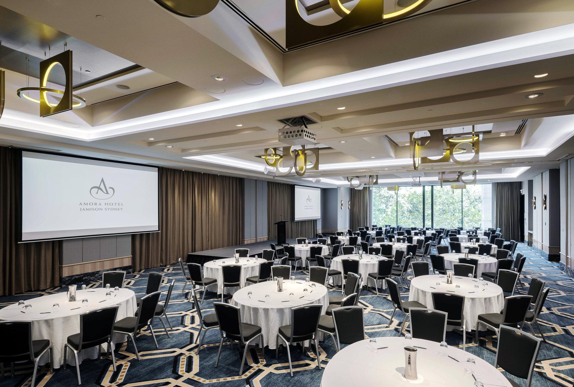 Amora Hotel Jamison Sydney Kültér fotó The photo shows a spacious conference or event room. There are multiple round tables arranged with white tablecloths and black chairs around them. The room has a modern design with elegant lighting fixtures hanging from the ceiling. A large screen is