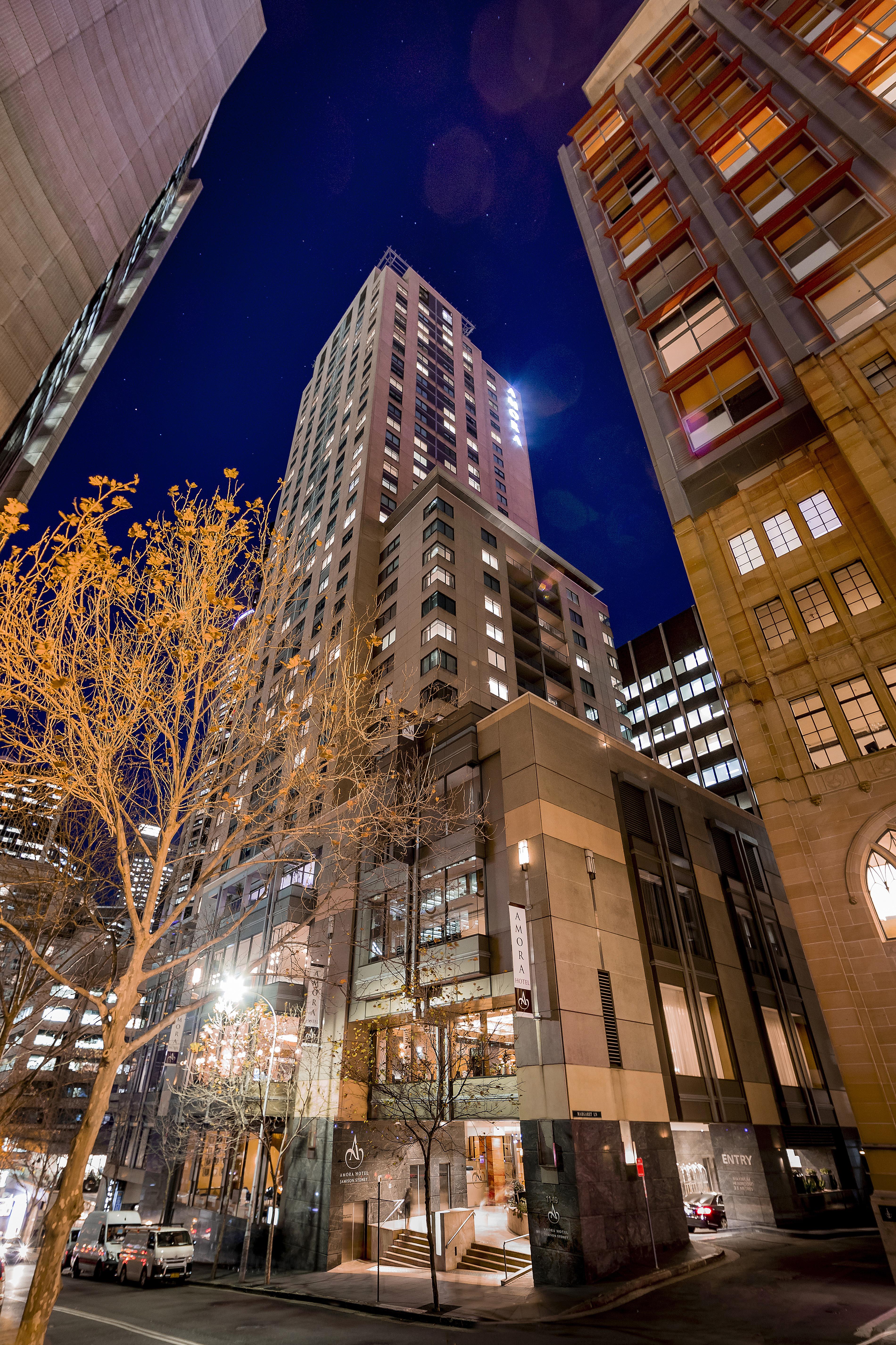 Amora Hotel Jamison Sydney Kültér fotó The photo shows a tall building illuminated at night, surrounded by other structures in an urban environment. The foreground features a tree with bare branches, suggesting it's possibly autumn or winter. The building itself has a modern design, with 