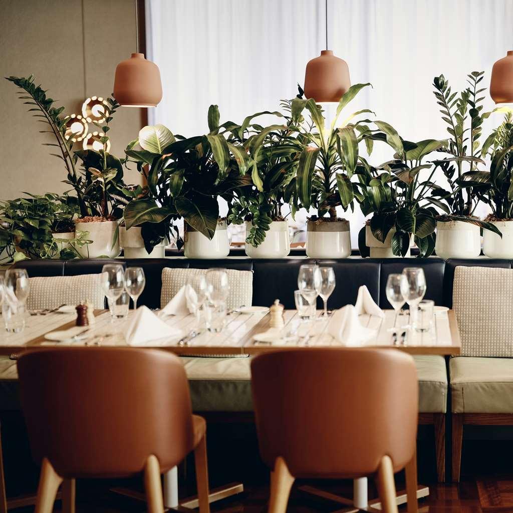 Amora Hotel Jamison Sydney Étterem fotó The photo shows a stylish restaurant interior featuring a warm and inviting atmosphere. There are neatly arranged tables with wooden surfaces, each set with glasses, napkins, and seasonings. Surrounding the tables are large, green plants in white pot