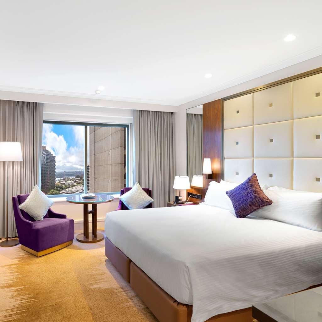 Amora Hotel Jamison Sydney Szoba fotó The photo shows a modern hotel room featuring a king-sized bed with white linens and a decorative purple pillow. To the side, there are two stylish armchairs in a deep purple color, accompanied by a small round table. The room has large windows cover