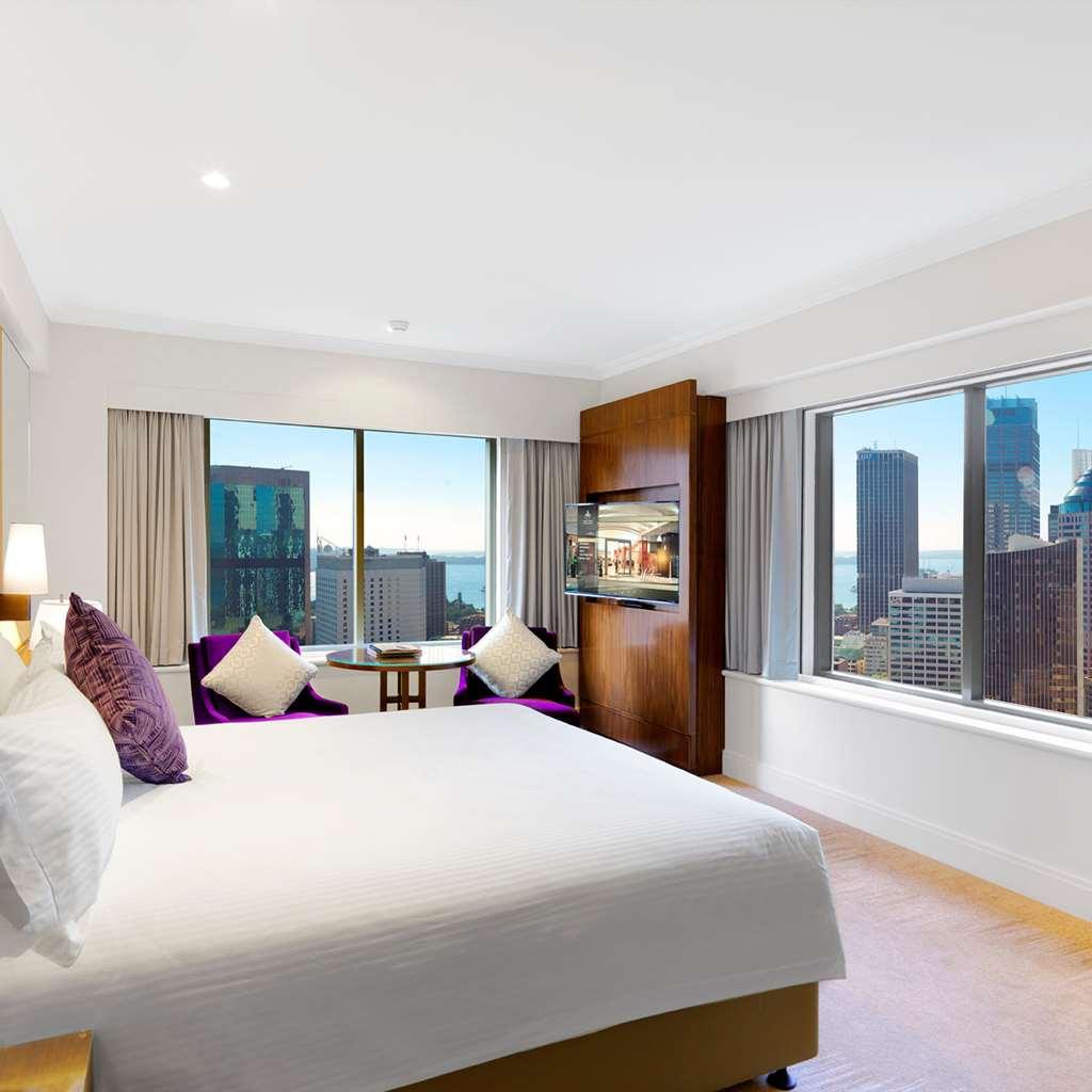 Amora Hotel Jamison Sydney Szoba fotó The photo shows a modern hotel room with large windows offering a view of a city skyline. The room features a large bed with white linens and purple accent pillows. There are two plush purple chairs next to a small round table by the window. A wooden