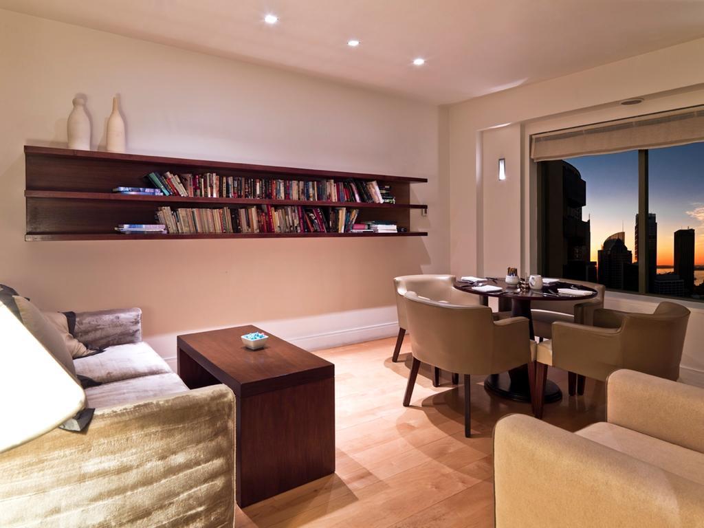 Amora Hotel Jamison Sydney Kültér fotó The photo shows a modern living space that features a cozy seating area. On the left, there is a sofa and armchair in neutral tones, complemented by a wooden coffee table in the center. A wall-mounted shelf holds a collection of books. To the right, 
