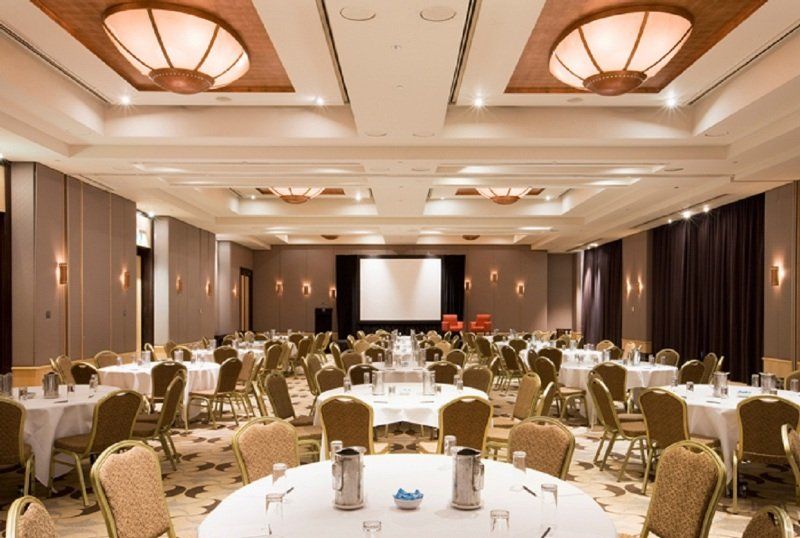 Amora Hotel Jamison Sydney Kültér fotó The photo shows a large banquet or conference room. There are several round tables arranged throughout the space, each covered with white tablecloths. The tables are set with water pitchers and glasses. In the center of the room, there is a projectio