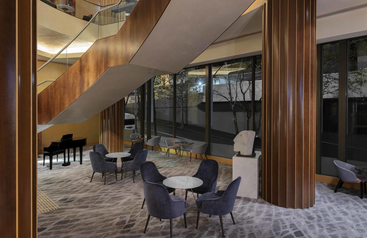 Amora Hotel Jamison Sydney Kültér fotó The photo depicts a modern interior space featuring a stylish lounge area. There are several comfortable chairs and small tables arranged on a patterned carpet. A large staircase with a sleek design is visible, leading to an upper level. The area is 