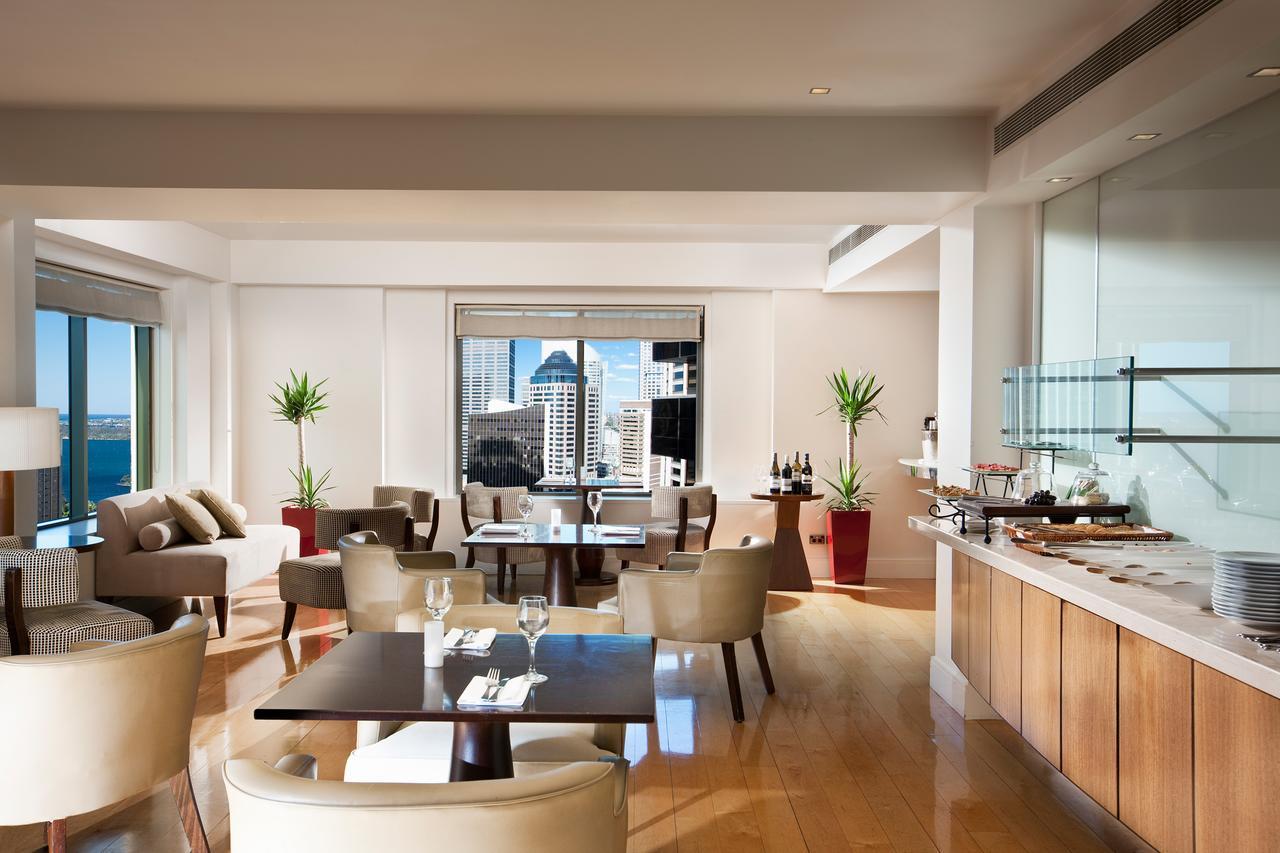 Amora Hotel Jamison Sydney Kültér fotó The photo depicts a modern and elegantly furnished dining area or lounge with large windows showcasing a cityscape view. The space features light-colored wooden floors and neutral-toned furnishings, including comfortable chairs and tables set for din
