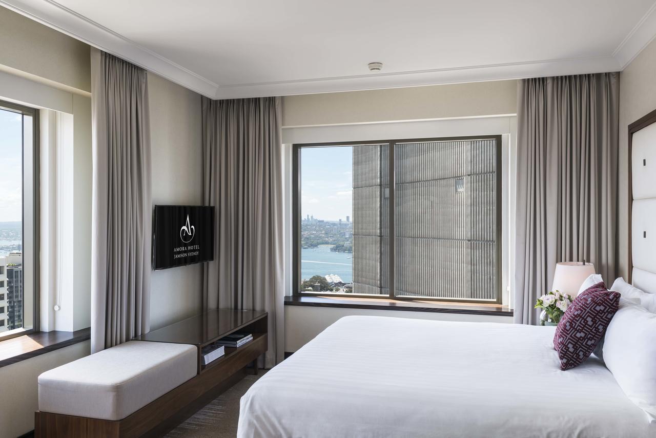 Amora Hotel Jamison Sydney Kültér fotó The photo shows a modern hotel room featuring a large bed with white bedding. There is a seating area with a bench next to a window. The room is well-lit with natural light coming in through the window, which has curtains. A flat-screen TV is mounted
