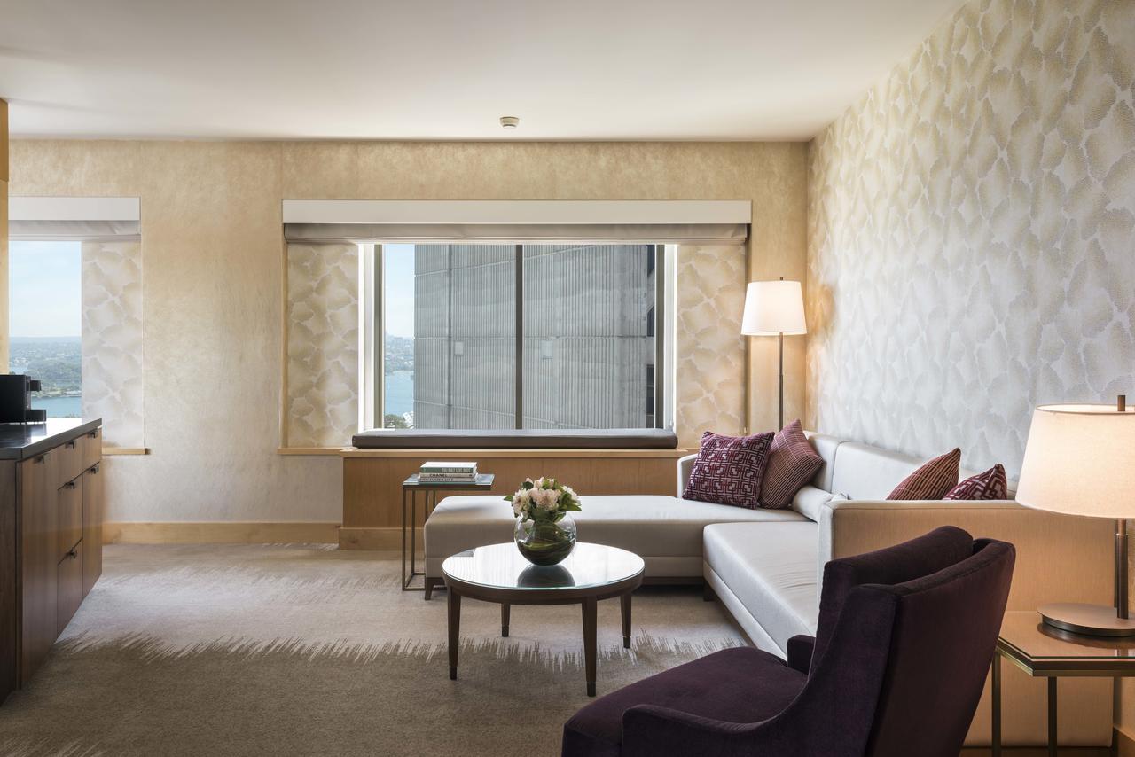 Amora Hotel Jamison Sydney Kültér fotó The photo shows a modern and stylish hotel room or living space. It features a large window with city views and light-colored walls adorned with a subtle pattern. There is a comfortable white sofa accompanied by purple accent pillows, positioned near