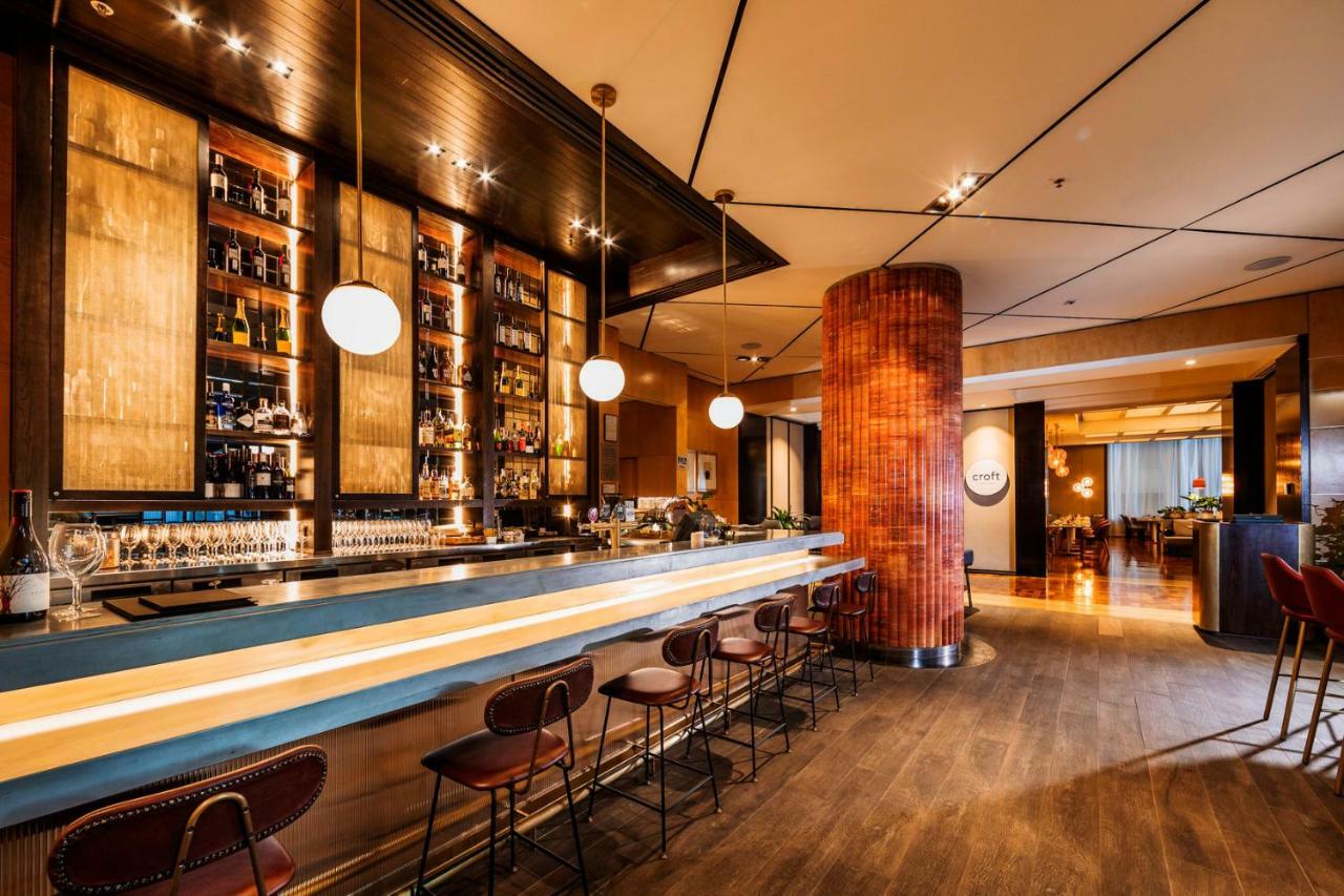 Amora Hotel Jamison Sydney Kültér fotó The photo shows the interior of a stylish bar. There is a long bar counter with a smooth surface, and several high-backed chairs lined up along it. Behind the bar, there is a wall of shelves filled with various types of bottles, likely containing spi
