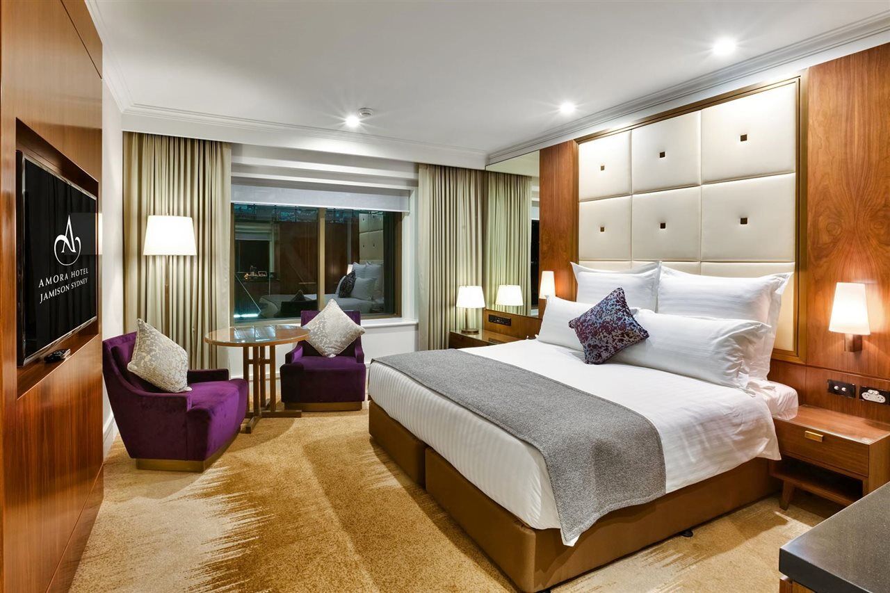 Amora Hotel Jamison Sydney Kültér fotó The photo shows a modern hotel room with a clean and stylish design. It features a large bed with white linens and a gray throw blanket. There are decorative pillows on the bed. To one side, there is a small seating area with two purple chairs and a 