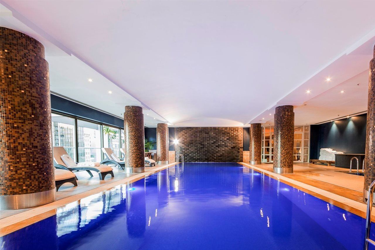 Amora Hotel Jamison Sydney Kültér fotó The photo features an indoor swimming pool with vibrant blue water. The pool area is well-lit, with several sources of light illuminating the space. Surrounding the pool are stylish stone columns, and there are lounge chairs positioned nearby for rel